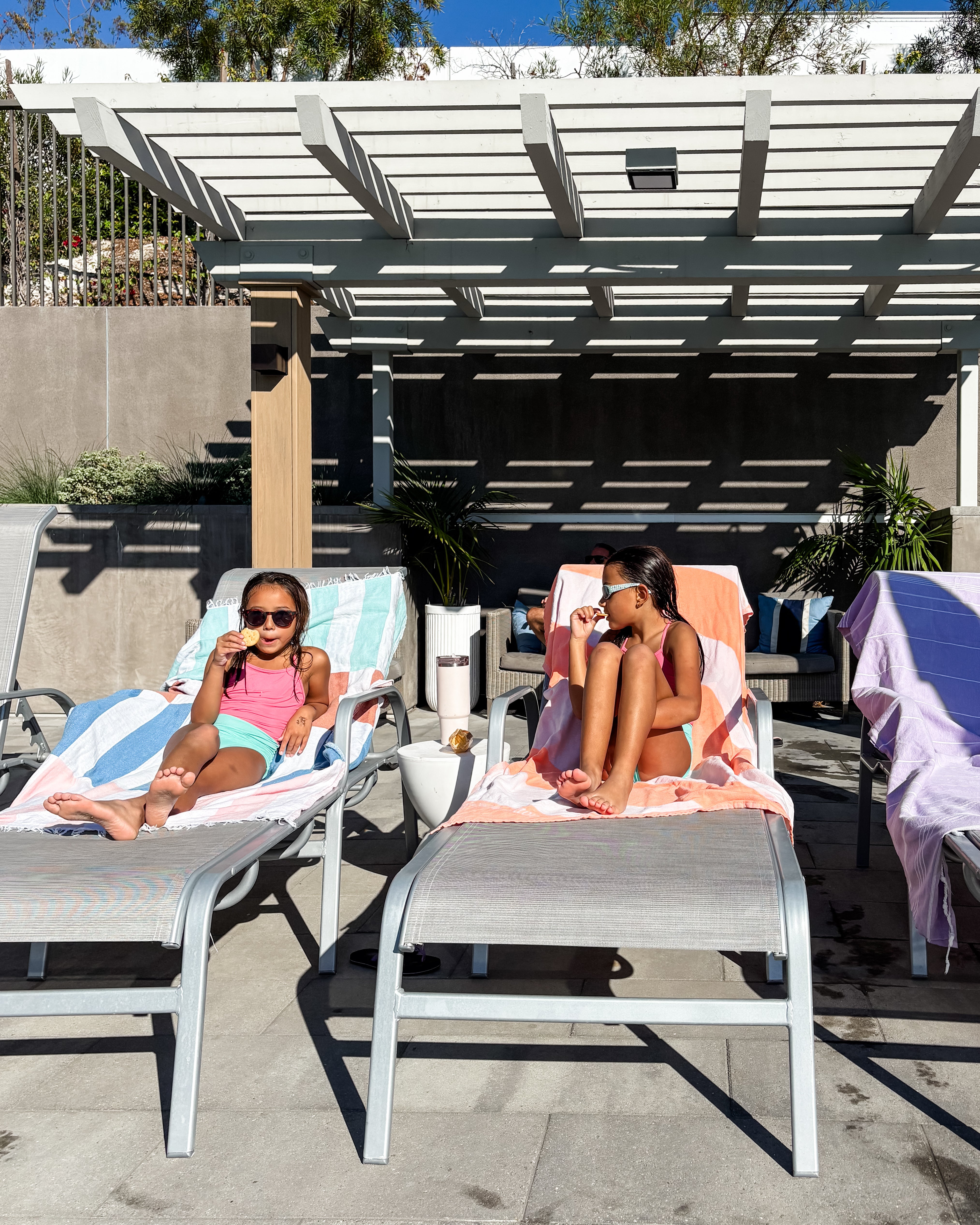 Zwei junge Mädchen in Badeanzügen entspannen sich am Pool und genießen ein Eis auf Liegestühlen, die mit Maxi Strandtuch De La Mer von Main Design bezogen sind. Bunte Handtücher und helles Sonnenlicht sorgen für einen verspielten Touch unter der Pergola.