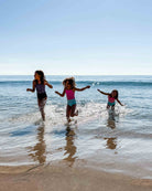 Drei junge Mädchen in dem Lena Mädchen Badeanzug mit UPF 50+ von Main Design spielen an einem sonnigen Tag im flachen Strandwasser. Die nachhaltigen Badeanzüge sind aus recycelten Materialien hergestellt, mit Spritzern um ihre Füße und dem Meer im Rücken.