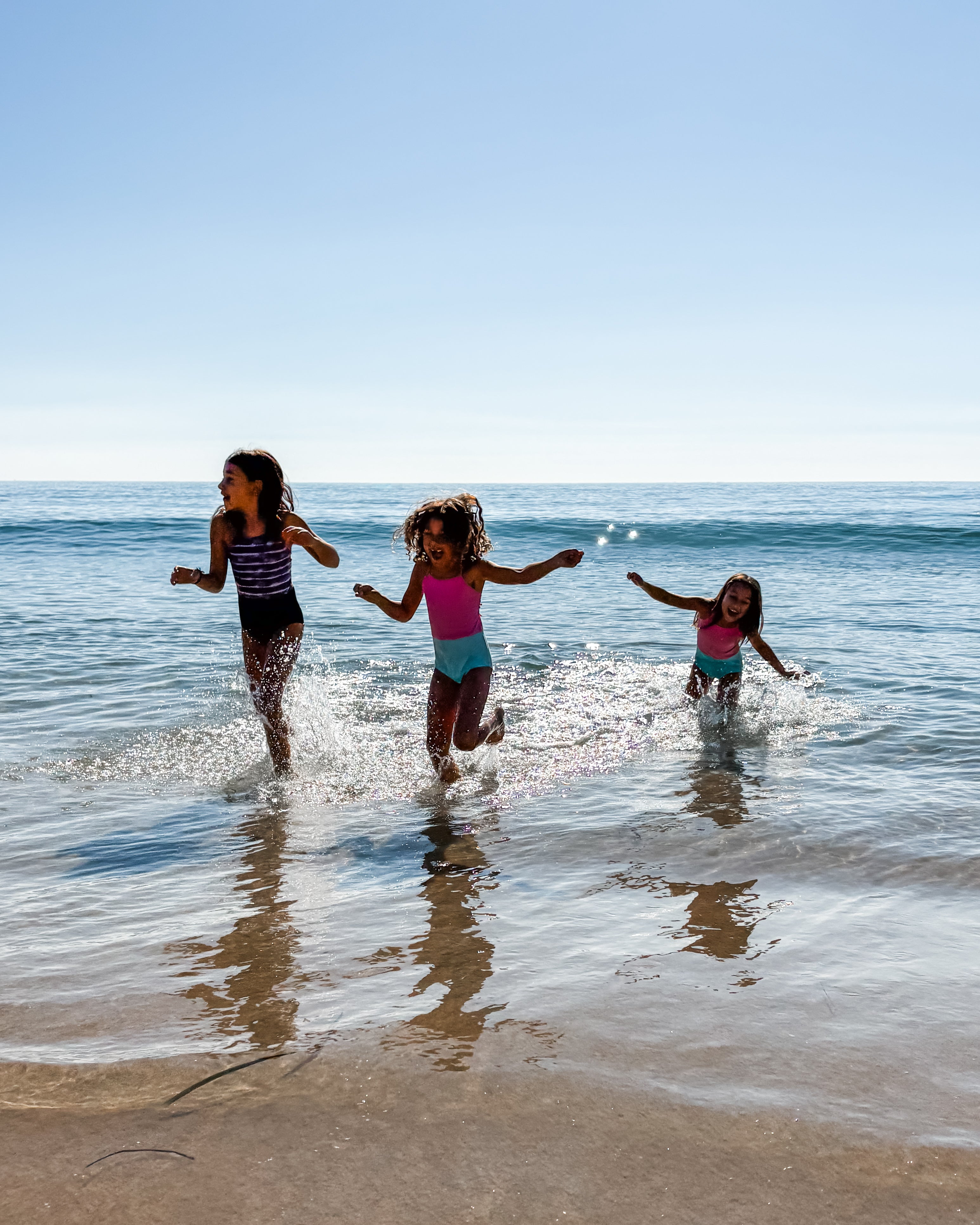 Drei junge Mädchen in dem Lena Mädchen Badeanzug mit UPF 50+ von Main Design spielen an einem sonnigen Tag im flachen Strandwasser. Die nachhaltigen Badeanzüge sind aus recycelten Materialien hergestellt, mit Spritzern um ihre Füße und dem Meer im Rücken.
