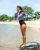 Eine Frau steht auf einem Reifen, der halb im seichten Wasser an einem Strand liegt. Sie trägt den Ivy Surf Suit von Main Design und eine schwarze Hose. Im Hintergrund sind ein Sandstrand, Bäume und ein klarer blauer Himmel zu sehen. Sie scheint ihre Haare oder Ohren zu richten.