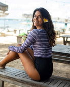 Eine Frau sitzt seitlich auf einer Holzbank am Strand und lächelt in die Kamera. Sie hat eine Blume im Haar und trägt einen Ivy Surf Suit von Main Design und schwarze Shorts aus recycelten Materialien. Der Strand ist mit Lichterketten dekoriert, im Hintergrund stehen Tische und Sonnenschirme.