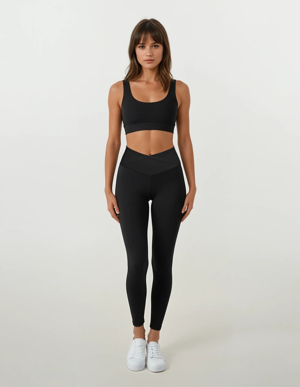Eine Frau blickt vor einem schlichten weißen Hintergrund nach vorne. Sie trägt die Juno Leggings von Main Design aus recyceltem Nylon mit hoher Taille, kombiniert mit einem schwarzen Sport-BH und weißen Turnschuhen.