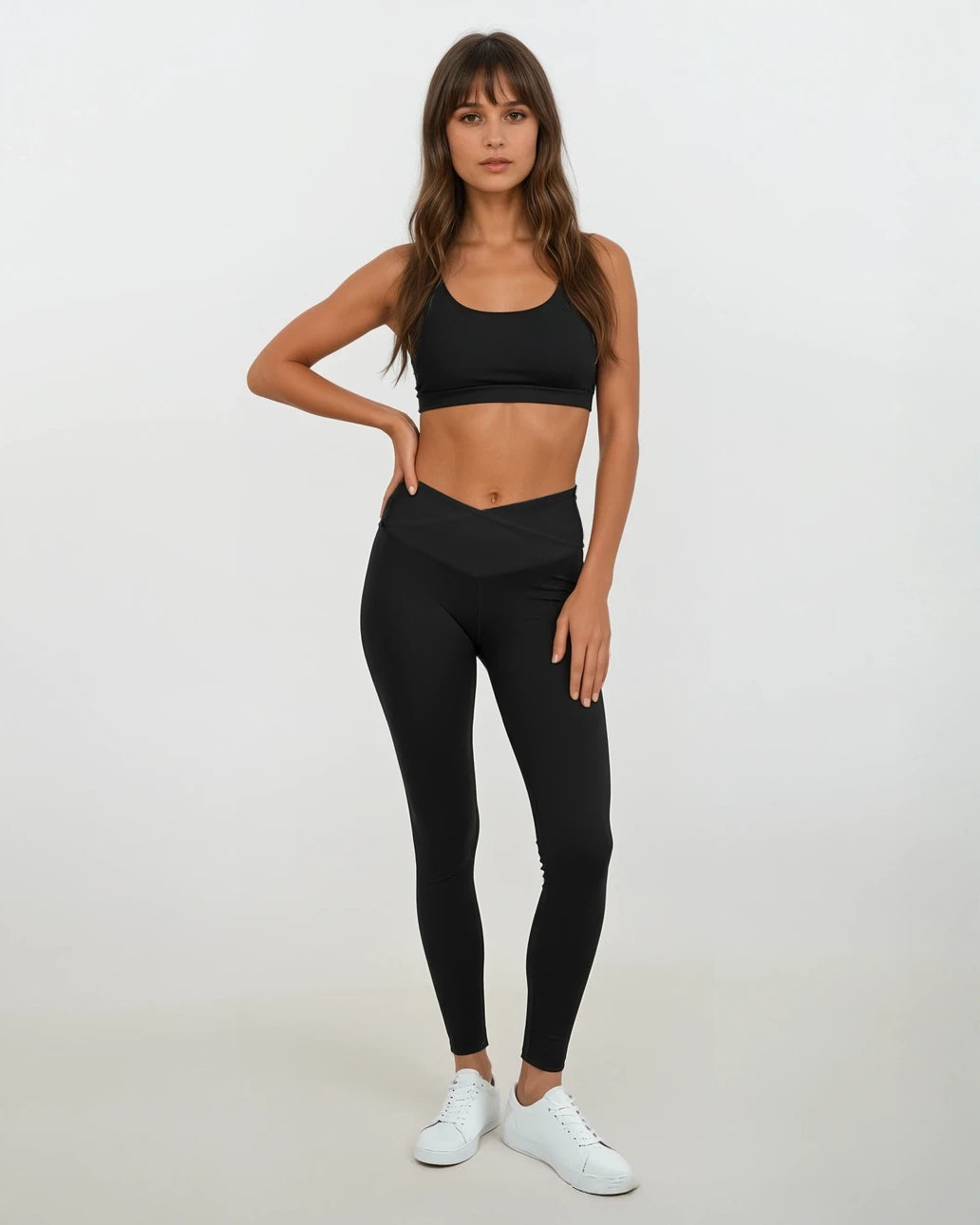 Eine Frau mit langen braunen Haaren trägt Main Design Juno Leggings mit hohem Bund aus recyceltem Nylon, einen schwarzen Sport-BH und weiße Sneakers vor weißem Hintergrund. Ihre linke Hand liegt auf der Hüfte.