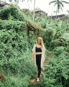Eine Frau mit langem blondem Haar geht barfuß über einen Steinweg inmitten üppiger tropischer Pflanzen und Ranken. Sie trägt den Kimono Saturn von Main Design, während im Hintergrund Palmen und Holzkonstruktionen zu sehen sind.