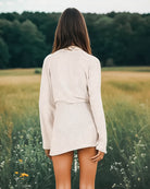 Eine Frau mit langen braunen Haaren steht mit dem Rücken zu einem Feld und trägt den Kimono Nadas von Main Design. Wildblumen umgeben sie, während sich unter einem bewölkten Himmel ein Wald abzeichnet.