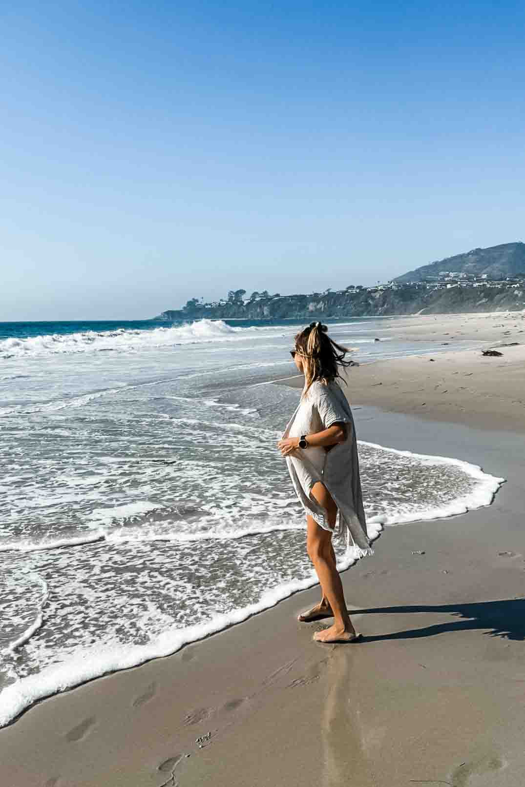 Eine Frau im Main Design Kimono Saturn aus nachhaltigen Materialien und Sandalen schlendert an einem Sandstrand entlang, während die Wellen anrollen. Der klare blaue Himmel gibt den Blick auf Hügel und ein paar Häuser in der Ferne frei.