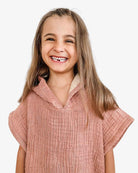 Ein lächelndes Kind mit langen braunen Haaren trägt den „Kinder Badeponcho Flow“ von Main Design aus OEKO-TEX zertifizierter Baumwolle. Der strukturierte rosa Musselinstoff verleiht dem reinweißen Hintergrund sanfte Eleganz.