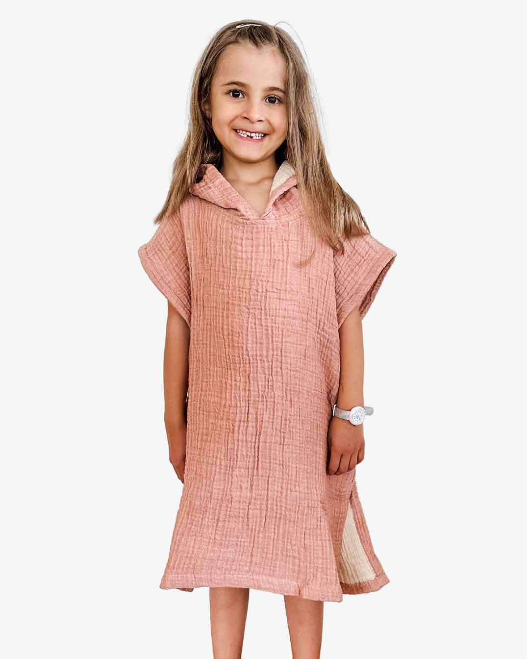 Ein junges Mädchen mit langen Haaren lächelt und trägt einen Main Design Kinder Badeponcho Flow in Pink. Sie ist aus OEKO-TEX zertifizierter Baumwolle gefertigt, steht barfuß vor einem schlichten weißen Hintergrund und trägt eine Uhr am linken Handgelenk.