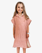 Ein junges Mädchen mit langen Haaren lächelt und trägt einen Main Design Kinder Badeponcho Flow in Pink. Sie ist aus OEKO-TEX zertifizierter Baumwolle gefertigt, steht barfuß vor einem schlichten weißen Hintergrund und trägt eine Uhr am linken Handgelenk.