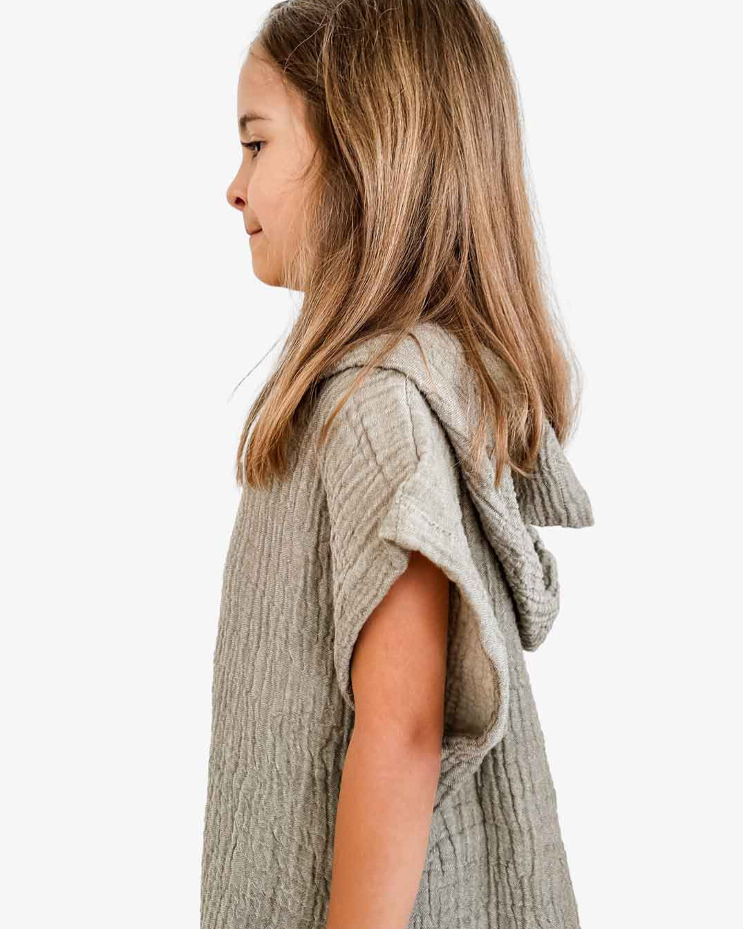 Ein junges Mädchen mit langen braunen Haaren steht im Profil, eingehüllt in einen Main Design Kinder Badeponcho Flow aus OEKO-TEX zertifizierter Baumwolle. Die hellgrüne Kapuze liegt über ihren Schultern, während der schlichte weiße Hintergrund die Textur des Musselinstoffs betont.