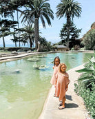 Zwei Kinder stehen an einem Pool und tragen passende hellrosa Kinder Badeponcho Flow-Outfits von Main Design, hergestellt aus OEKO-TEX zertifizierter Baumwolle. Der Pool liegt in einem üppigen Garten mit Palmen vor einem klaren blauen Himmel.