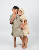 Zwei Kinder posieren vor einem schlichten Hintergrund. Der Junge, der ein albernes Gesicht macht, trägt einen grauen OEKO-TEX-zertifizierten Baumwollmantel, während das Mädchen in einem beigen Main Design Kinder Badeponcho Flow lächelt und seinen Arm hält. Beide sind barfuß und strahlen Freude und Unbeschwertheit aus.