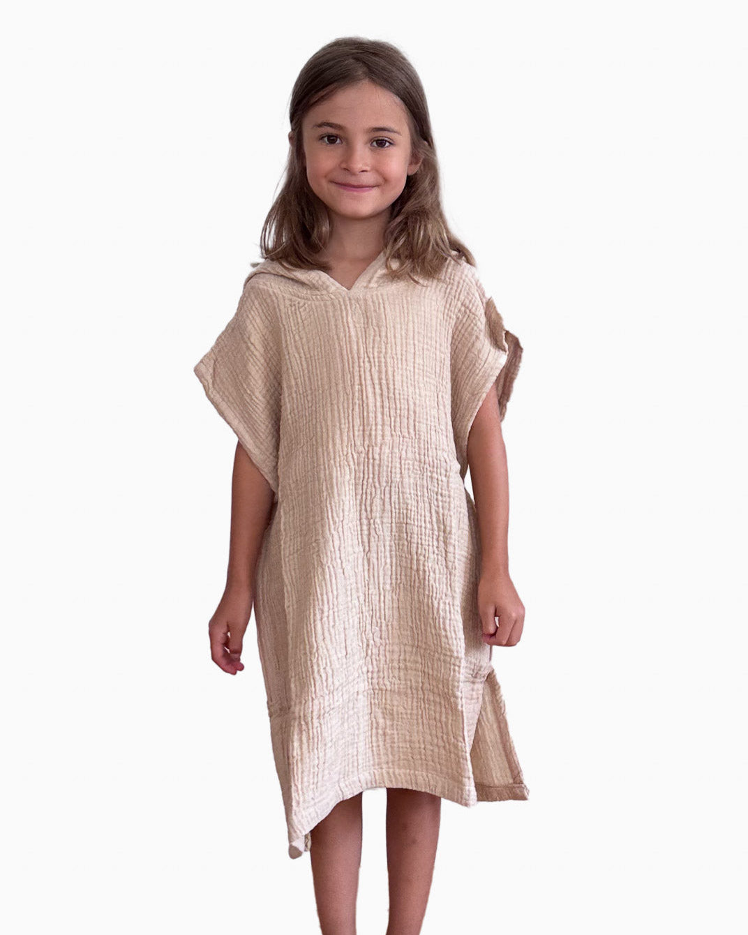 Ein junges Mädchen mit langen braunen Haaren steht barfuß und lächelt. Sie trägt ein locker sitzendes, strukturiertes beiges Kleid aus Musselin von Main Design aus OEKO-TEX-zertifizierter Baumwolle. Das Produkt ist der Kinder Badeponcho Flow, der vor einem schlichten weißen Hintergrund steht.