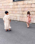 Zwei Kinder im Main Design Kinder Badeponcho Flow laufen barfuß auf einem gepflasterten Weg an einer Steinmauer. Das eine trägt einen leichten Poncho mit Sandalen, das andere ein rosafarbenes Musselin-Handtuch und ist barfuß - beide schauen beim Gehen nach unten.