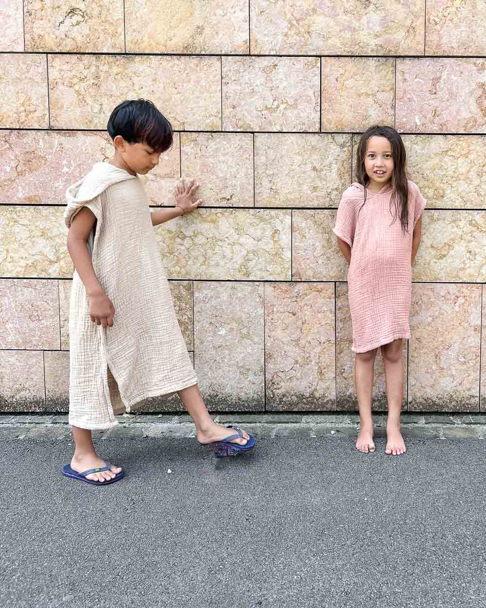 Zwei Kinder stehen vor einer hellbraunen Steinmauer. Der Junge links trägt einen hellbeigen Main Design Kinder Badeponcho Flow und blaue Sandalen, während das Mädchen rechts ein hellrosa Musselin-Kleid trägt und barfuß in die Kamera lächelt.