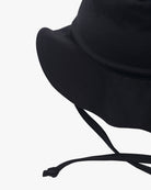 Eine Nahaufnahme eines schwarzen Kinder-Sonnenhuts von Main Design mit breiter Krempe und verstellbarem schwarzen Kinnriemen. Der Hut aus umweltfreundlichem Stoff ruht auf einem schlichten weißen Hintergrund und unterstreicht sein einfaches und funktionales Design mit zusätzlichem Sonnenschutz UPF 50+.