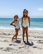 Zwei junge Mädchen im Ava Mädchen Badeanzug von Main Design, passend in Gelb und Hellblau, stehen an einem Sandstrand und lächeln in die Kamera, während im Hintergrund das Meer und der Himmel zu sehen sind.