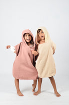 Zwei lächelnde Kinder stehen barfuß auf einem einfarbigen Hintergrund, gekleidet in den kuscheligen Main Design Kinder Badeponcho Flow. Das eine trägt Rosa, das andere Beige, aus weichem Musselin aus OEKO-TEX zertifizierter Baumwolle. Mit umeinander geschlungenen Armen nehmen sie eine verspielte Pose ein.