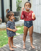 Zwei Kinder spielen draußen mit Seifenblasen. Ein Kind, das ein rotes Oberteil und schwarze gemusterte Shorts trägt, macht Seifenblasen. Das andere Kind, das einen schwarz-weiß gestreiften Laila Mädchen-Badeanzug von Main Design trägt, schaut zu und lächelt. Sie stehen auf einem Holzdeck mit Grün im Hintergrund.