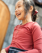 Ein junges Mädchen mit zurückgebundenen langen Haaren, das einen roten langärmeligen Laila Mädchen-Badeanzug von Main Design trägt, lächelt strahlend. Sie trägt eine Halskette und Ohrringe und scheint zu lachen. Der Hintergrund ist verschwommen und zeigt eine helle Ziegelwand und eine Holzverkleidung.