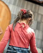 Eine Person mit nassen Haaren, die mit einem Haargummi zu einem Pferdeschwanz gebunden sind, trägt einen roten Laila Mädchen-Badeanzug von Main Design aus umweltfreundlichem Stoff mit LSF 50+ Sonnenschutz und ist von hinten aufgenommen. Sie steht vor einer Holztür und einer strukturierten Steinmauer, was darauf hindeutet, dass sie sich im Freien befindet.