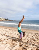 Ein Kind, das den Laila Mädchen Badeanzug von Main Design aus recycelten Materialien mit Sonnenschutzfaktor 50+ trägt, macht einen Handstand an einem Sandstrand mit Meer und Klippen im Hintergrund unter einem teilweise bewölkten Himmel.
