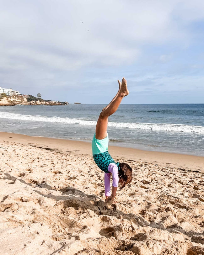 Ein Kind, das den Laila Mädchen Badeanzug von Main Design aus recycelten Materialien mit Sonnenschutzfaktor 50+ trägt, macht einen Handstand an einem Sandstrand mit Meer und Klippen im Hintergrund unter einem teilweise bewölkten Himmel.