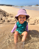 Ein kleines Kind mit einem lilafarbenen Sonnenhut und dem türkisfarbenen Leopardenprint Laila Mädchen Badeanzug von Main Design sitzt lächelnd im flachen Wasser an einem sonnigen Sandstrand.