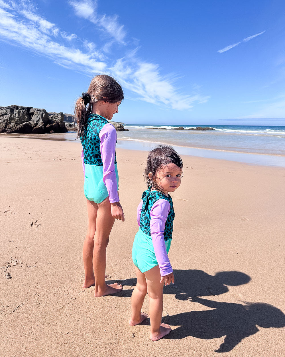 Zwei junge Mädchen im aqua- und lilafarbenen Laila Mädchen Badeanzug von Main Design stehen barfuß an einem Sandstrand und blicken unter einem klaren Himmel auf das Meer, mit sanften Wellen und Felsen im Hintergrund.
