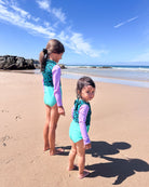Zwei junge Mädchen im aqua- und lilafarbenen Laila Mädchen Badeanzug von Main Design stehen barfuß an einem Sandstrand und blicken unter einem klaren Himmel auf das Meer, mit sanften Wellen und Felsen im Hintergrund.