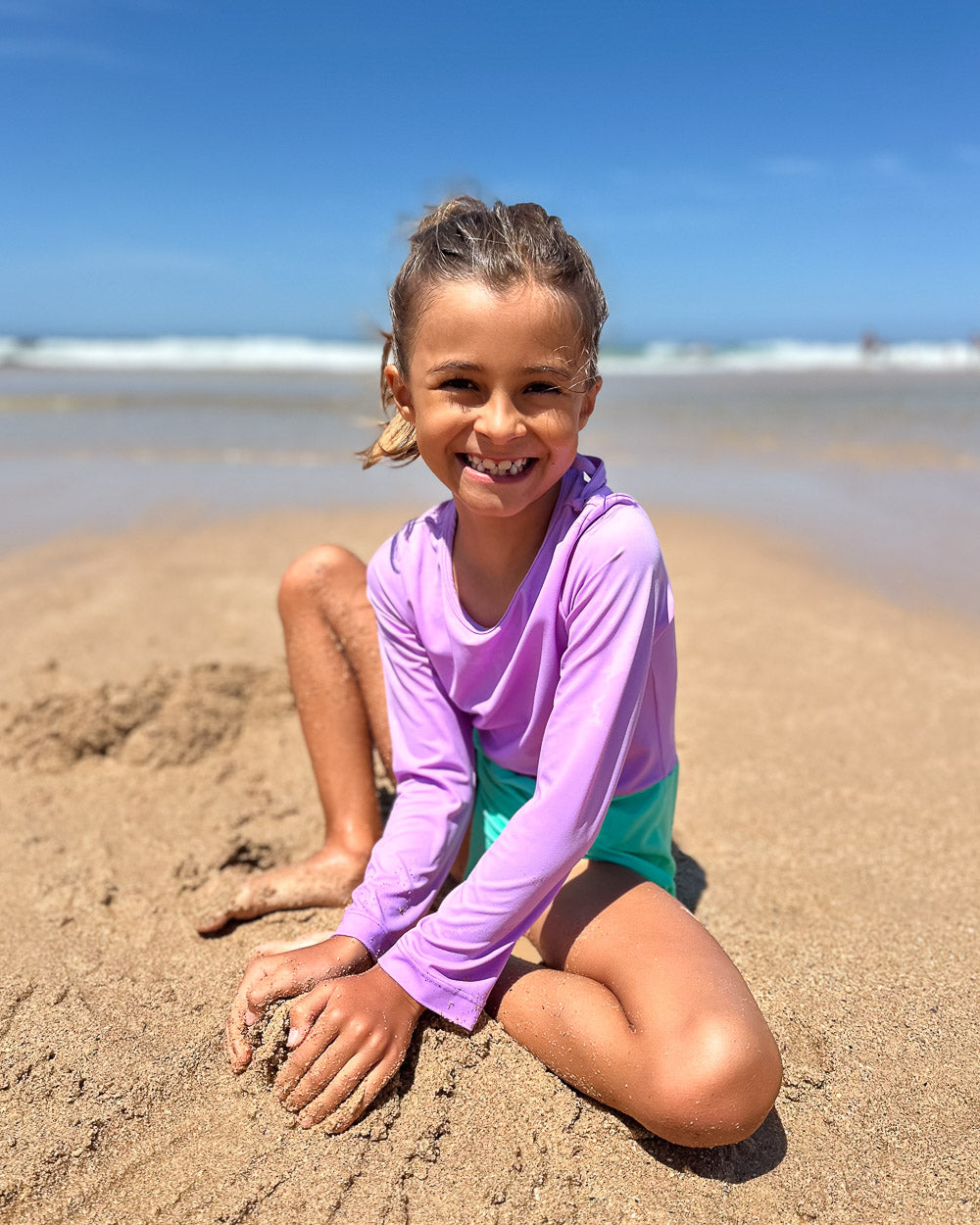 Ein lächelndes Kind, das den Main Design Laila Mädchen Badeanzug und die blaugrünen Shorts trägt, genießt den Strand, das Meer und den blauen Himmel - und bleibt dabei mit UPF 50+ geschützt.