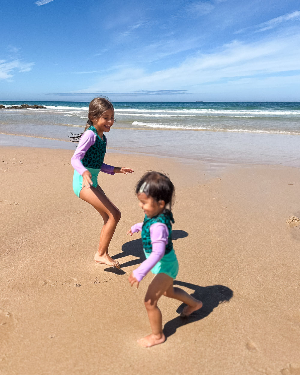 Zwei kleine Kinder im passenden Main Design Laila Mädchen Badeanzug mit UPF 50+ laufen und spielen an einem sonnigen Strand in der Nähe des Wassers, umgeben von sanften Wellen und einem klaren blauen Himmel.