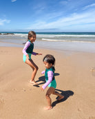 Zwei kleine Kinder im passenden Main Design Laila Mädchen Badeanzug mit UPF 50+ laufen und spielen an einem sonnigen Strand in der Nähe des Wassers, umgeben von sanften Wellen und einem klaren blauen Himmel.