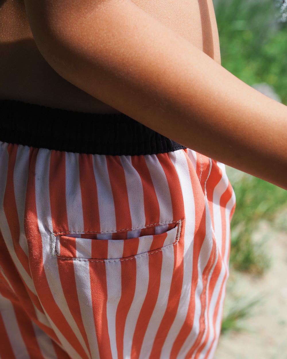 Ein Kind trägt Main Design Lean Boardshorts für Jungen in rot-weiß gestreift mit einem schwarzen Bund. Die schnell trocknenden Shorts bieten UPF 50+ und sind im Freien abgebildet, mit grünen Pflanzen, die im Hintergrund sanft verschwimmen.