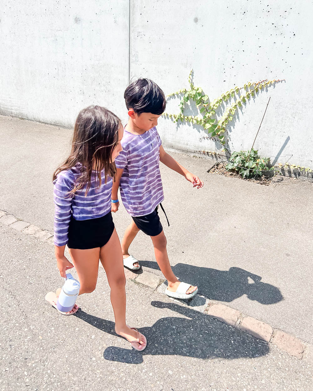 Zwei Kinder in passenden lila gestreiften Hemden und schwarzen Shorts gehen auf einem sonnigen Bürgersteig spazieren. Das Mädchen, das den Main Design Laila Mädchen Badeanzug mit UPF 50+ trägt, hält eine Wasserflasche, während sich ihre Schatten an einer mit Weinreben bewachsenen Mauer erstrecken.