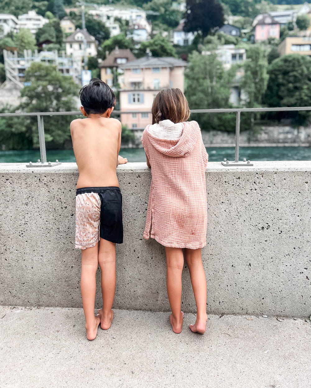 Zwei Kinder stehen barfuß an einem Betonzaun und blicken auf den Fluss und die entfernten Häuser. Das eine trägt eine Badehose, das andere ist in einen weichen Main Design Kinder Badeponcho Flow aus Musselin OEKO-TEX Baumwolle gehüllt.