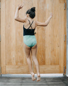 Ein junges Mädchen in einem schwarz-grünen Badeanzug mit verstellbaren Trägern springt mit dem Rücken zur Kamera. Ihr Haar ist zu einem Knoten zusammengebunden und sie schwebt in der Luft vor großen Holztüren. Der Holzboden unter ihr zeigt einen kleinen nassen Bereich von ihrem schnell trocknenden, nachhaltigen Lena Mädchen Badeanzug von Main Design.