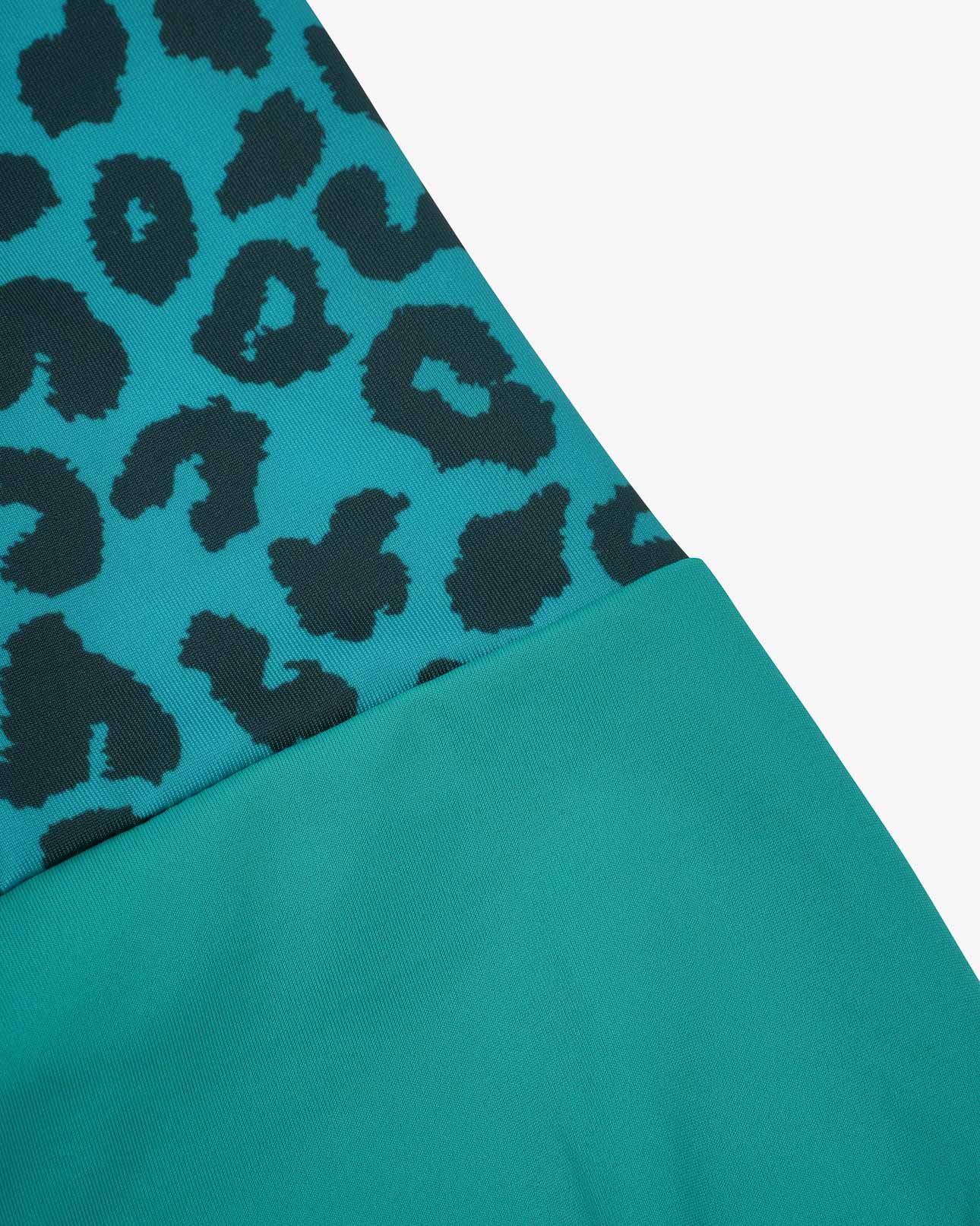 Nahaufnahme eines türkisfarbenen Lena Mädchen-Badeanzugs von Main Design mit Leopardenmuster aus recycelten Materialien. Der Stoff hat eine glatte Textur mit unregelmäßigen schwarzen Formen auf einer Seite und einfarbigem Türkis auf der anderen, wodurch ein stilvoller diagonaler Übergang entsteht.