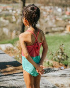 Ein junges Mädchen mit nassen Haaren steht im Freien in einem pink- und blaugrünen Lena Mädchen Badeanzug von Main Design aus recycelten Materialien und blickt mit hinter dem Rücken verschränkten Händen vor einem grasbewachsenen, unscharfen Hintergrund in die Ferne.