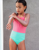 Ein junges Mädchen trägt den Lena Mädchen Badeanzug von Main Design in Rosa und Mintgrün aus recycelten Materialien, während sie sich mit nassen Haaren den Arm eincremt. Sie steht vor einer grauen Wand und schaut konzentriert nach unten.