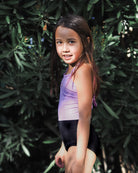 Ein junges Mädchen mit langen braunen Haaren steht im Freien vor grünen Büschen, trägt den hellvioletten Lena Mädchen Badeanzug von Main Design aus recycelten Materialien und lächelt sanft, während das Sonnenlicht durch die Blätter fällt.