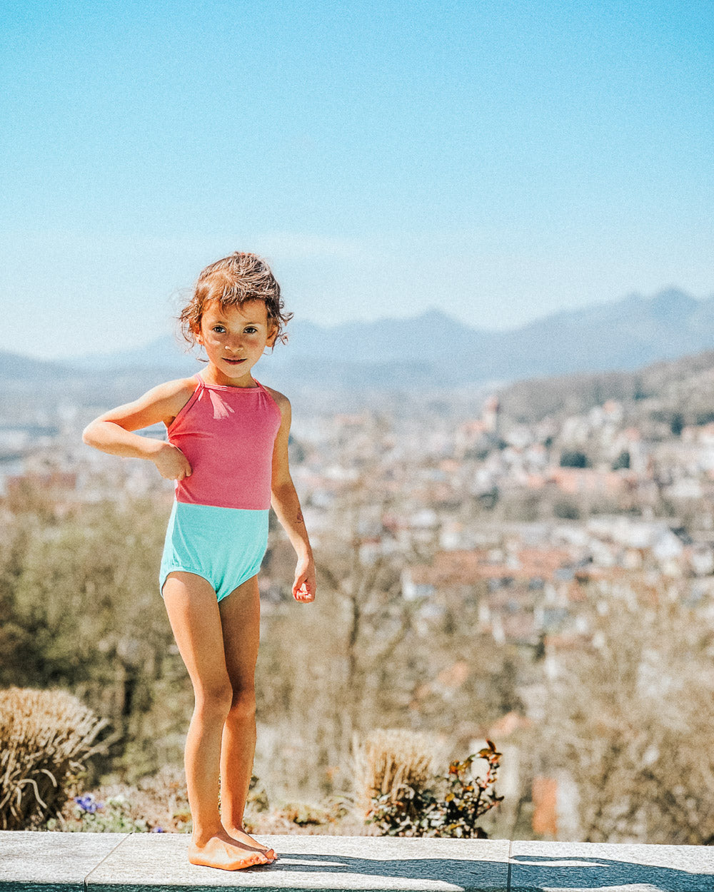 Ein kleines Kind trägt den Lena Mädchen Badeanzug von Main Design, einen roten und hellblauen UPF 50+-Badeanzug, und posiert im Freien auf einem Stein mit einer verschwommenen Stadtlandschaft und Bergen unter einem klaren blauen Himmel im Hintergrund.