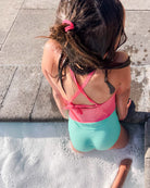 Ein junges Mädchen mit nassen Haaren und einem rosafarbenen Haargummi sitzt am Beckenrand, trägt den Lena Mädchen Badeanzug von Main Design in Rosa und Mintgrün und taucht ihre Füße ins Wasser, während das Sonnenlicht Schatten auf die Fliesen wirft.