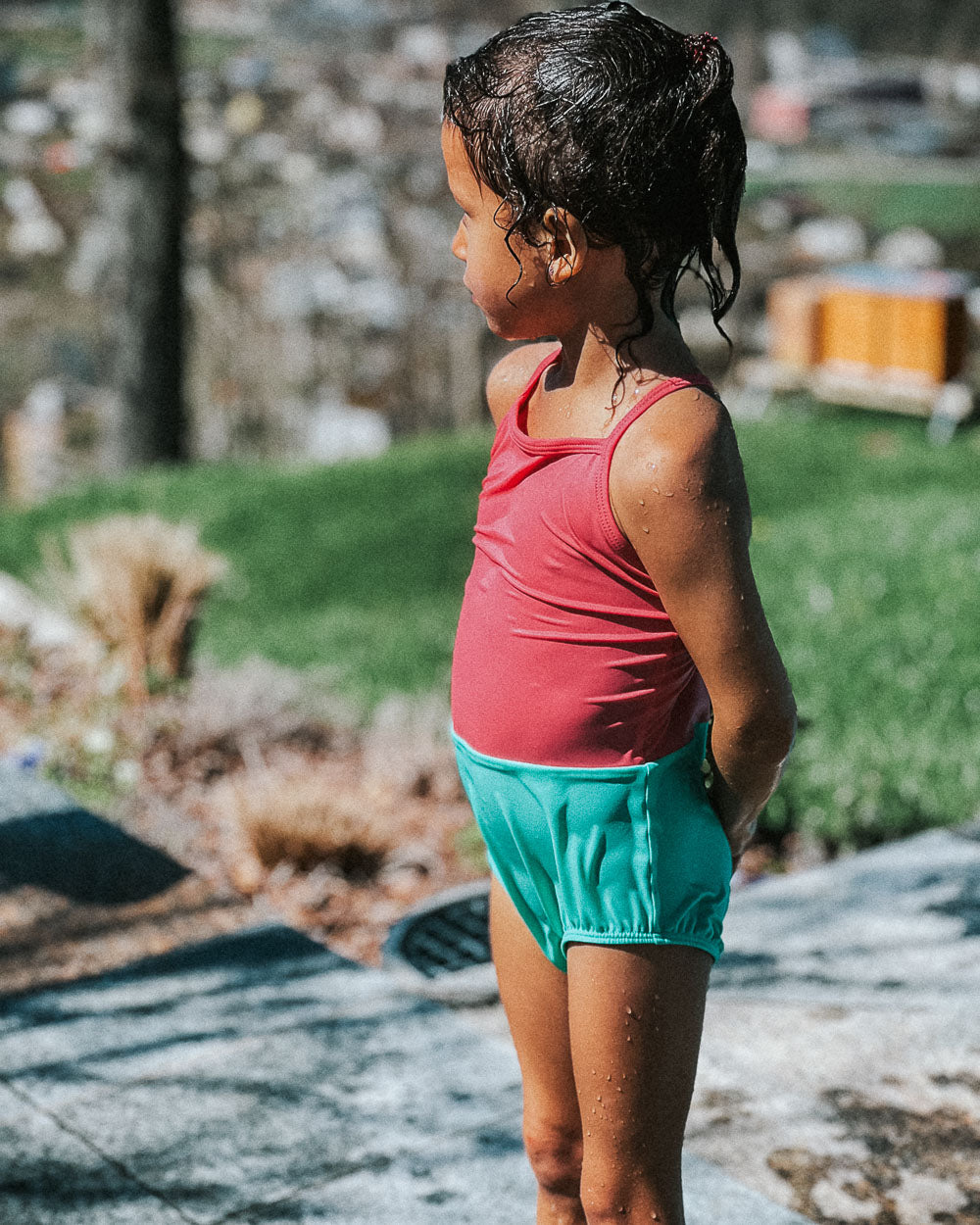 Ein kleines Kind im Main Design Lena Mädchen Badeanzug - mit rotem UPF 50+ Oberteil und blaugrünen Shorts - steht mit nassen Haaren auf einem sonnenbeschienenen, grasbewachsenen Bürgersteig und genießt den Tag in diesem nachhaltigen Badeanzug.