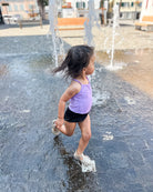 Ein kleines Kind in einem lilafarbenen Lena Mädchen Badeanzug von Main Design und schwarzen Shorts läuft an einem sonnigen Tag fröhlich durch einen Wasserbrunnen auf einem gepflasterten öffentlichen Platz.