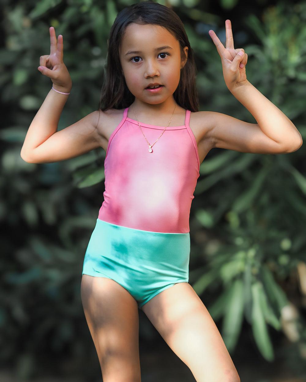 Ein Mädchen trägt den Lena Mädchen Badeanzug von Main Design in Pink und Türkis, steht vor grünem Hintergrund, zeigt das Peace-Zeichen und blickt in die Kamera.