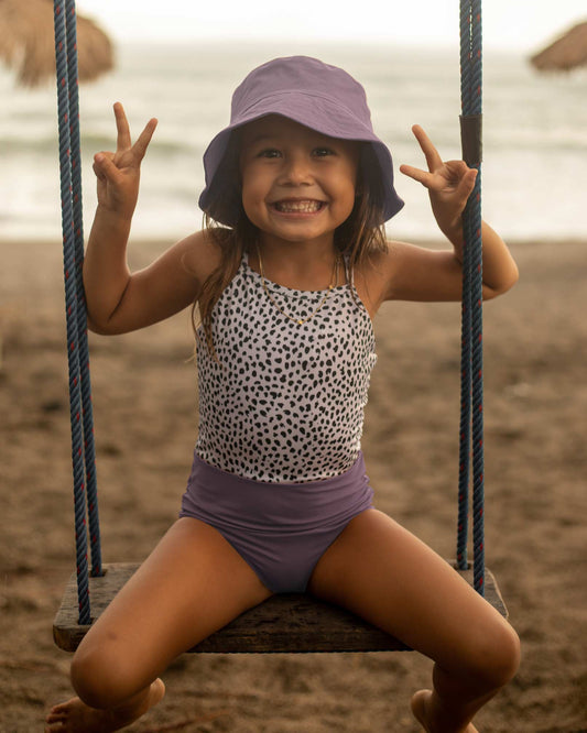 Ein junges Mädchen in einem Main Design Lena Mädchen Badeanzug und einem lila Fischerhut sitzt breit lächelnd auf einer Schaukel und zeigt mit beiden Händen das Peace-Zeichen. Der Sandstrand und das Meer im Hintergrund unterstreichen ihre Fröhlichkeit.