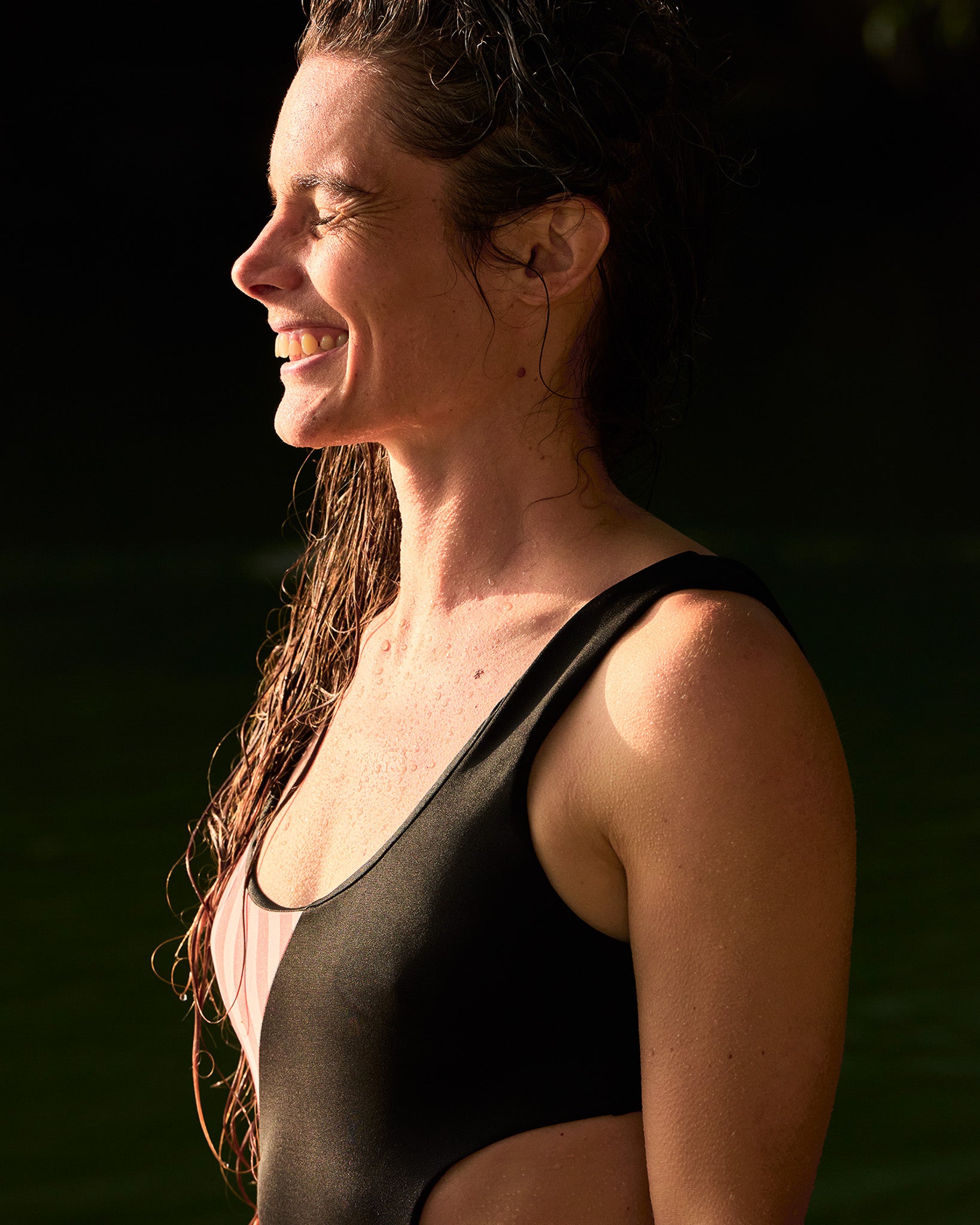 Eine Frau mit nassen Haaren lächelt im Sonnenlicht, trägt den Main Design Luna Badeanzug mit seitlichen Cut-outs und steht im Freien am Wasser.
