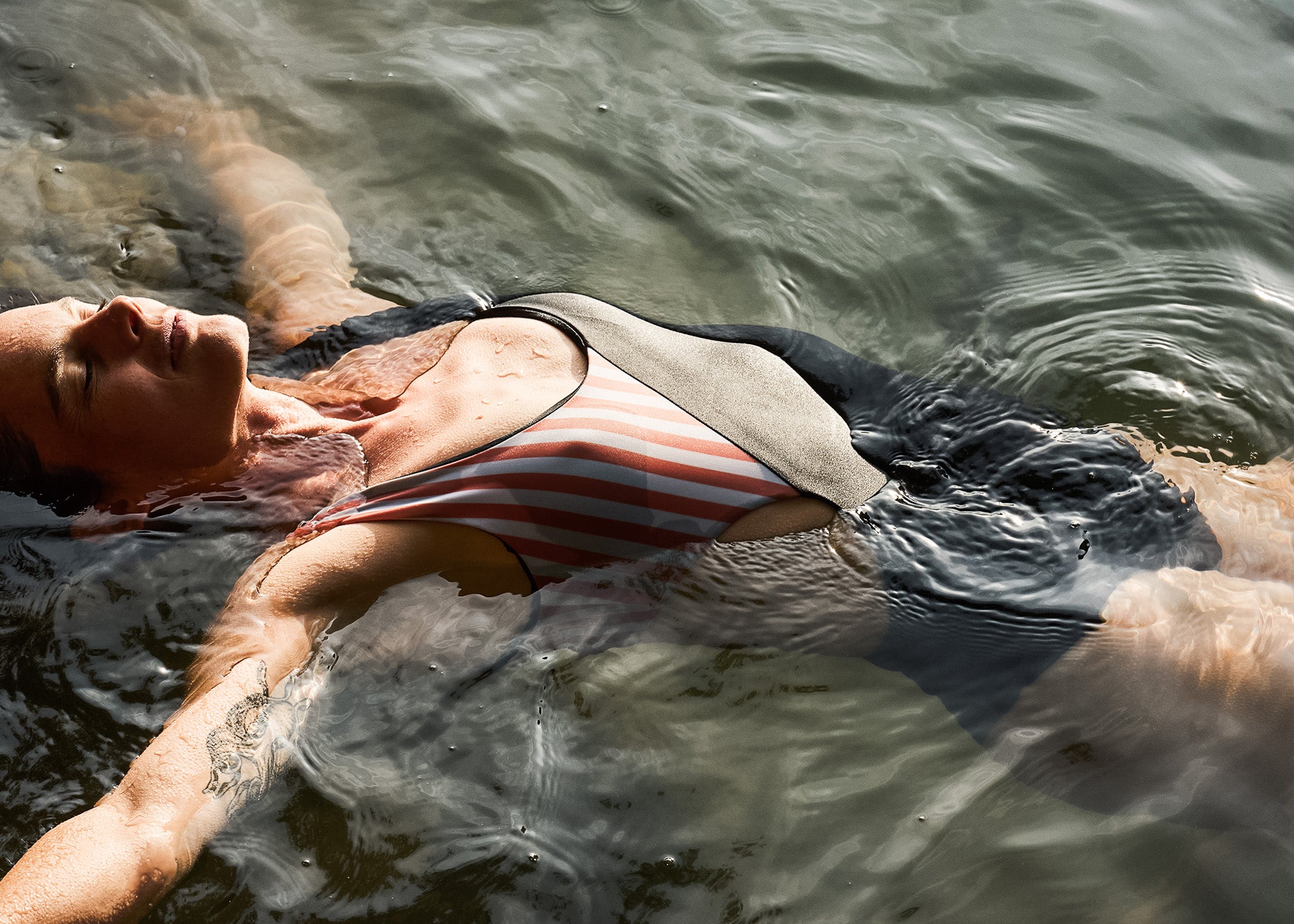 Der Main Design Luna Badeanzug mit seitlichen Ausschnitten zeigt eine Person, die mit ausgestreckten Armen und geschlossenen Augen auf dem Rücken im sonnenbeschienenen Wasser schwebt und teilweise untergetaucht ist.