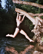 Eine Frau im Badeanzug Luna von Main Design, mit seitlichen Ausschnitten und aus recycelten Materialien gefertigt, hängt lächelnd von einem Ast über dem Wasser. Im Hintergrund sind üppiges grünes Laub und schimmerndes Wasser zu sehen.